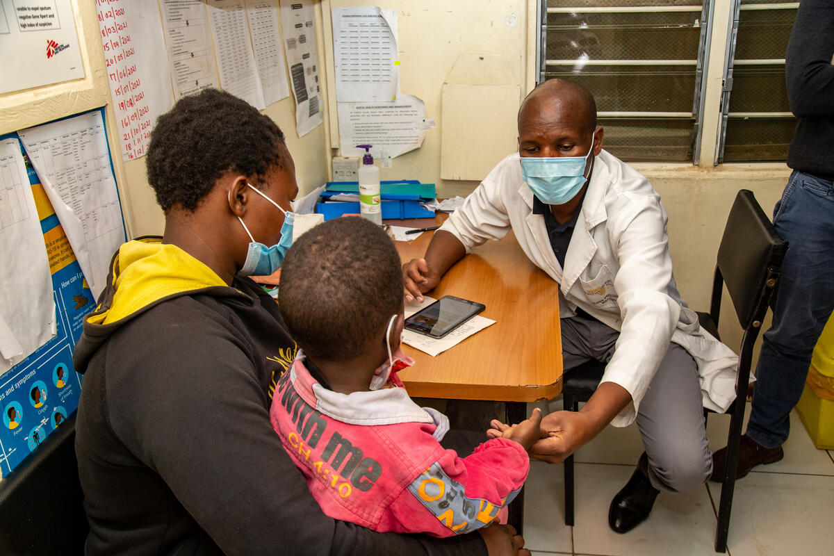 A doctor, wearing a mask and sitting at a desk, consults with a mother and child