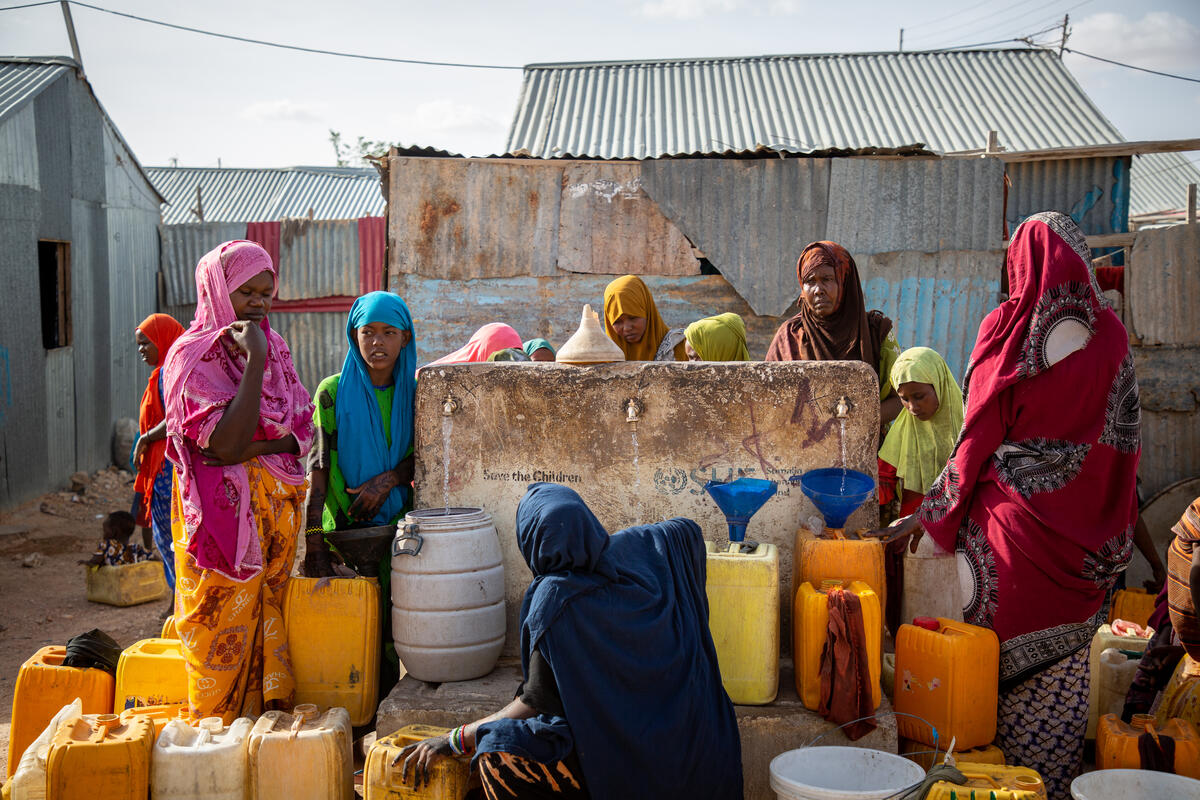 A group of women gather around a well and water source filling bidons of water.