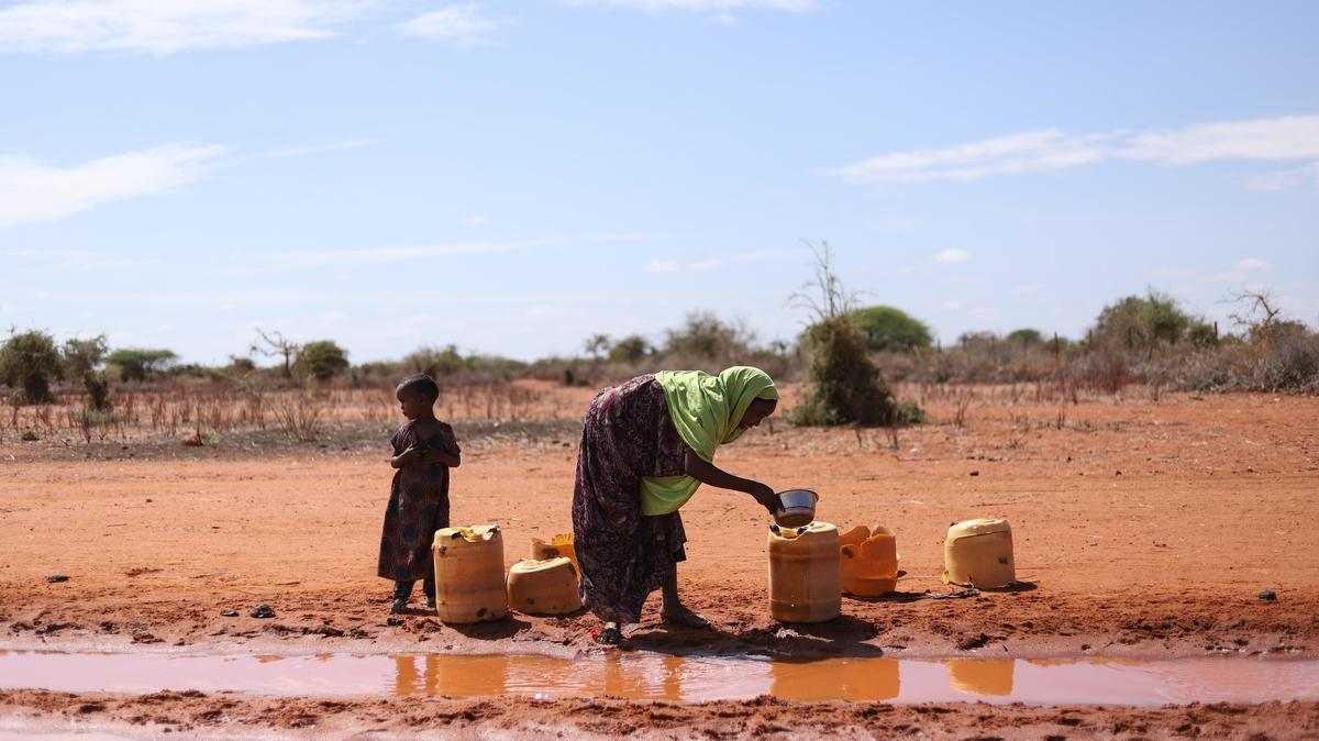A woman fetching water from a puddle in Kenya, pouring it into yellow bidons. There is a small child accompanying her.