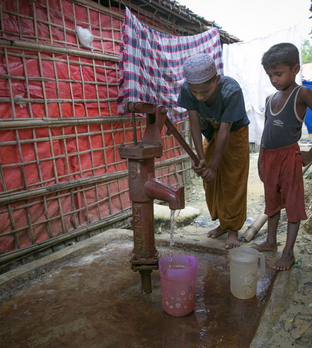 Two young boys pump water from a well in between two buildings.