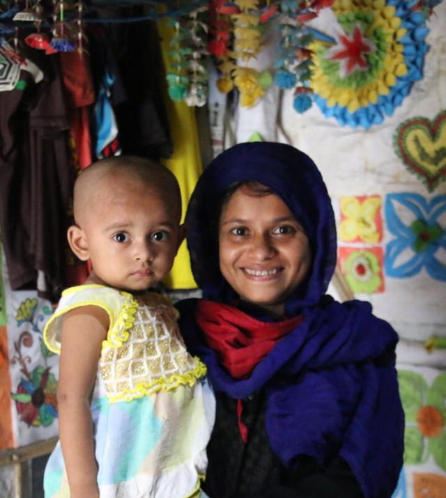 A smiling mother wearing a blue headscarf holder her young child in her arms.