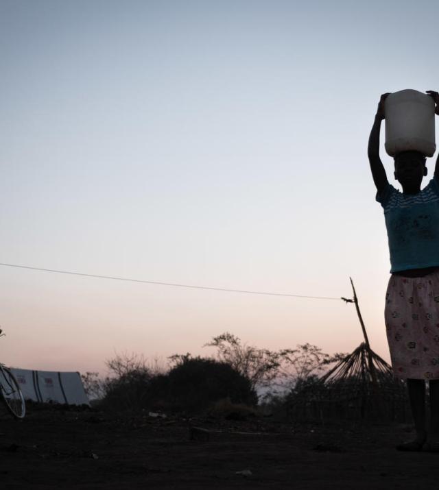 A young girl carries a jerry can of water on her head, the photo is in shadow with a sunset in the background.