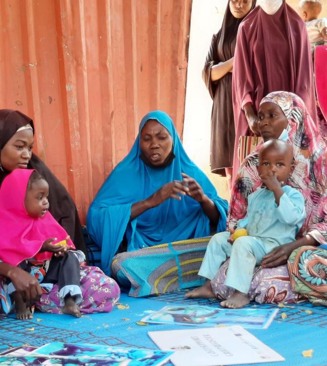Four women holding two small children sit on a blue map in discussion.