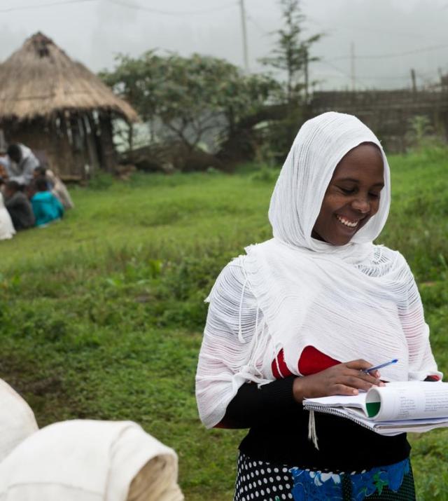 A woman wearing a white headscarf holds a notepad and smiles.