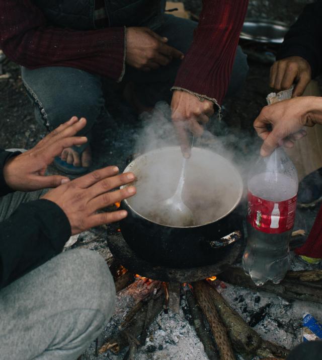 A group of people with their hands outstretched crouch around a pot that is boiling over a wood fire.