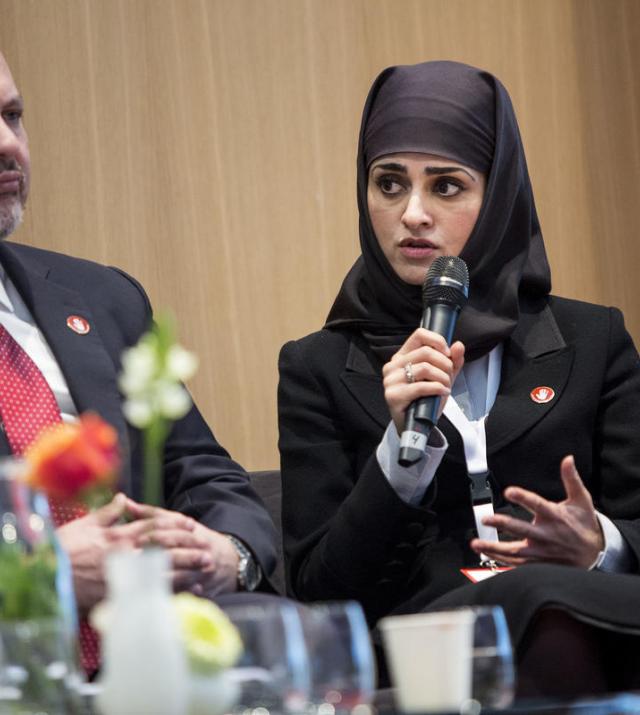 A woman wearing a black headscarf sits next to a man. She is speaking into a microphone and gesturing with her hand.