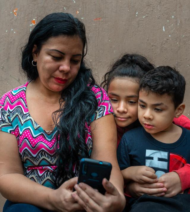 Une mère tient un smartphone à la main tandis que ses deux jeunes enfants le regardent par-dessus son épaule.