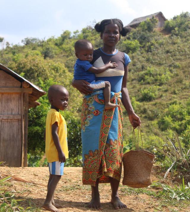 A woman carries a child on her hip and a basket in her other hand while standing next to a young child outside in Madagascar countryside.