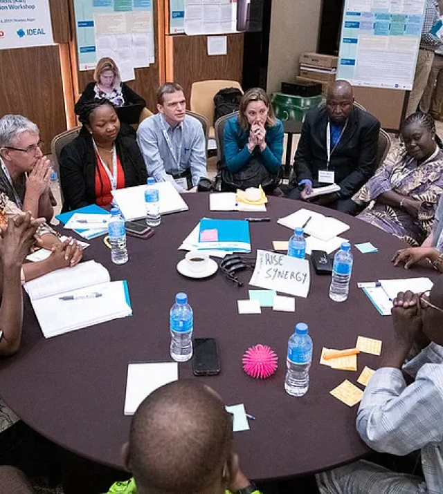 A group of men and women sit around a table at a conference. They are in discussion and thinking.