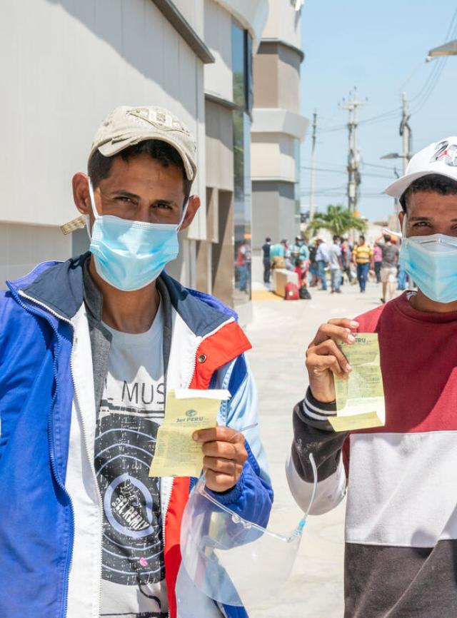 Two men wearing facemasks and baseball hats hold up cash assistance vouchers, and each raise a fist with a thumbs up