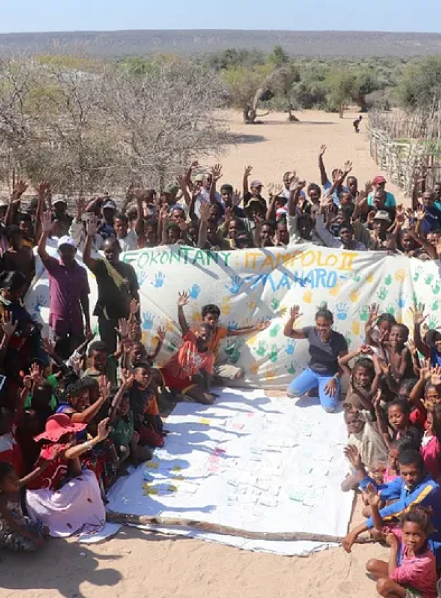 Un groupe d’enfants et d’adultes lèvent la main et se rassemblent autour d’un drap sur lequel sont gravées des empreintes de mains et du texte peint.