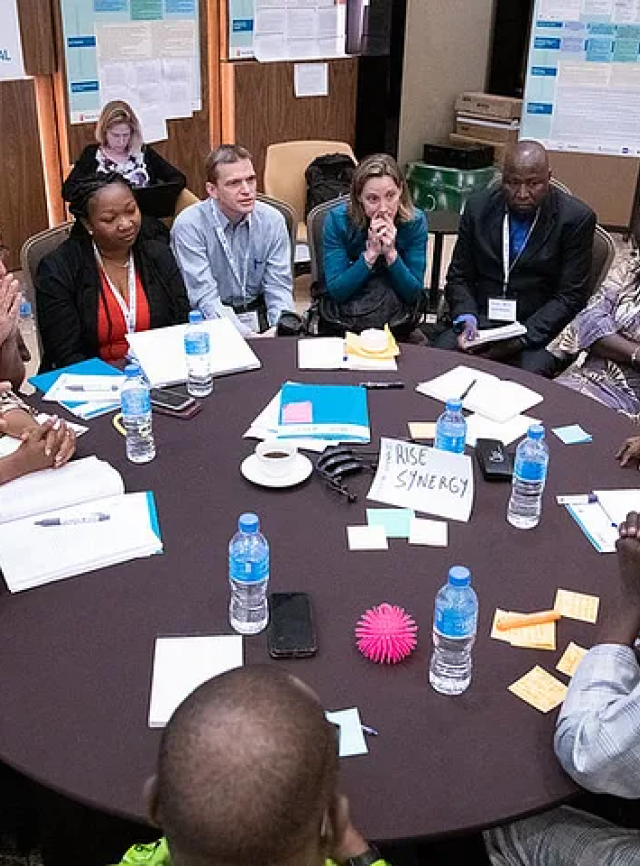 A group of men and women sit around a table at a conference. They are in discussion and thinking.