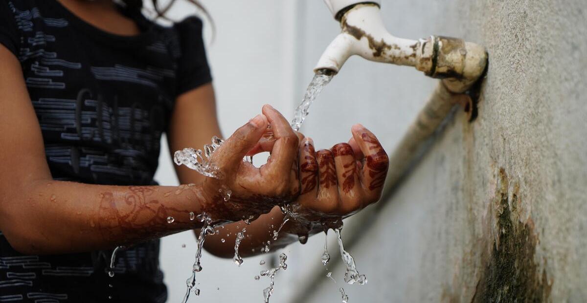 Young girl washes hands from water spout on a wall.