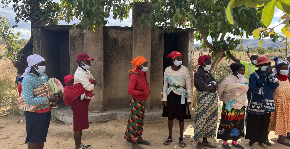 A group of women wearing face masks line up outside a latrine.