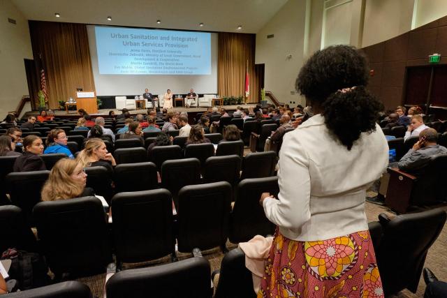 Photo of a woman standing at the back of an auditorium facing the stage to ask a question during a conference