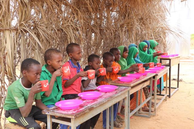 A group of children sit at their desks drinking tea awaiting a meal to be served.