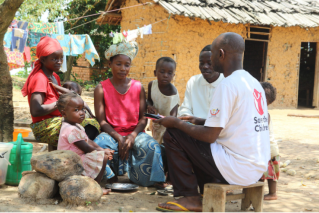 A Save the Children staff member sits and talks with a man, a woman, and a group of children.