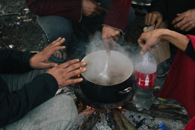 A group of people with their hands outstretched crouch around a pot that is boiling over a wood fire.