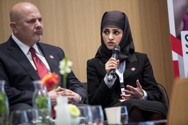 A woman wearing a black headscarf sits next to a man. She is speaking into a microphone and gesturing with her hand.