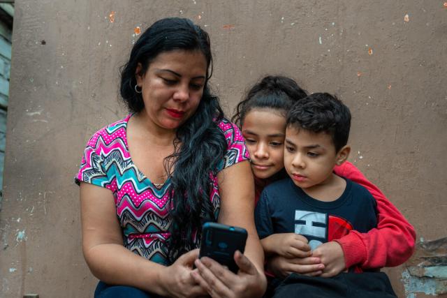 A mother holds a smartphone in her hand as her two young children look over her shoulder at it.